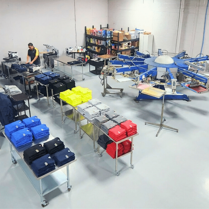Commercial screen printing facility with a multi-station press and tables of folded shirts in various colors.
