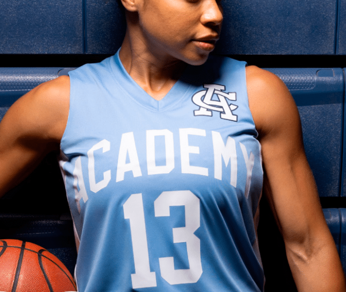 A person in a light blue "Academy 13" basketball jersey holding a basketball against a dark blue background.