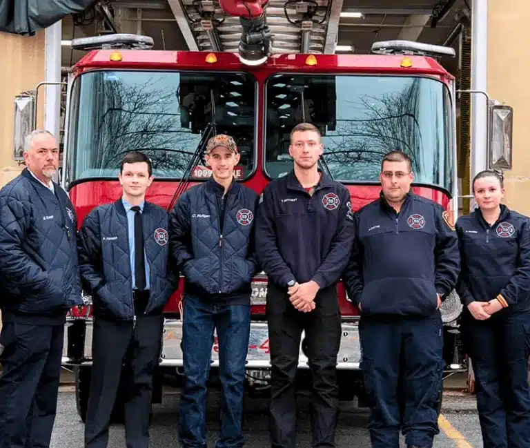 A group of six firefighters in uniform standing in front of a red fire engine.