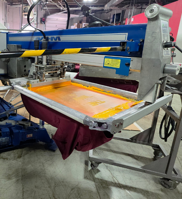 Maroon T-shirt being screen printed with yellow ink on a commercial screen printing press.